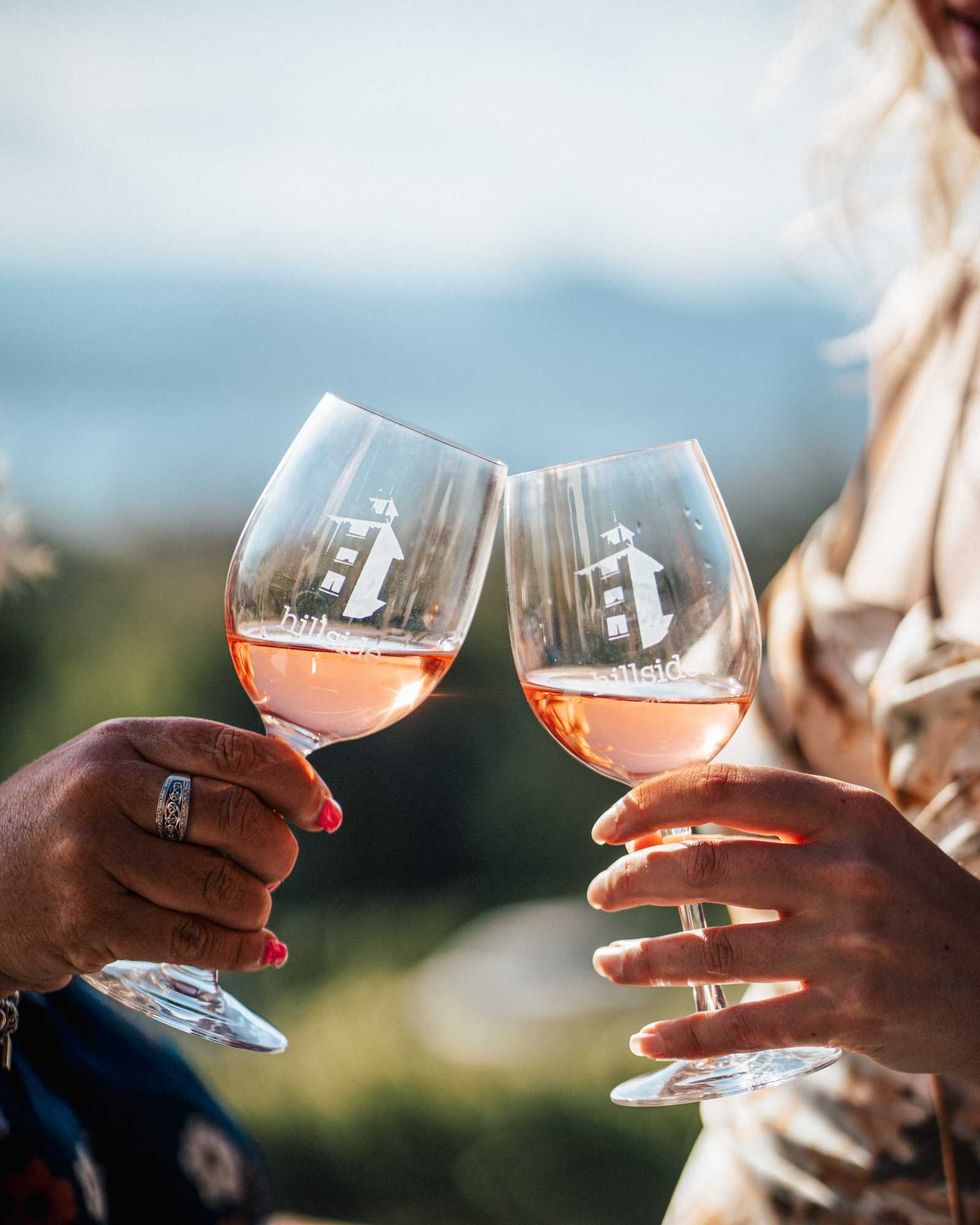 Two people cheers glasses of ros\u00e9 with the logo for Hillside Winery on the glasses.