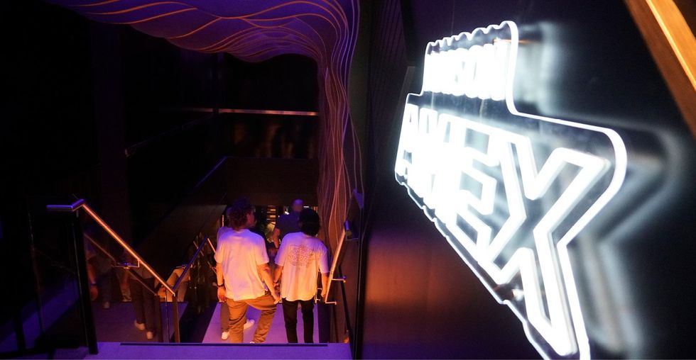 Two people in white shirts walk down the stairs next to a neon sign that reads "Maison Amex."