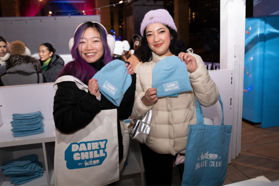 Two people smile as they hold up toques bearing the "MilkUp Dairy Chill" logo.