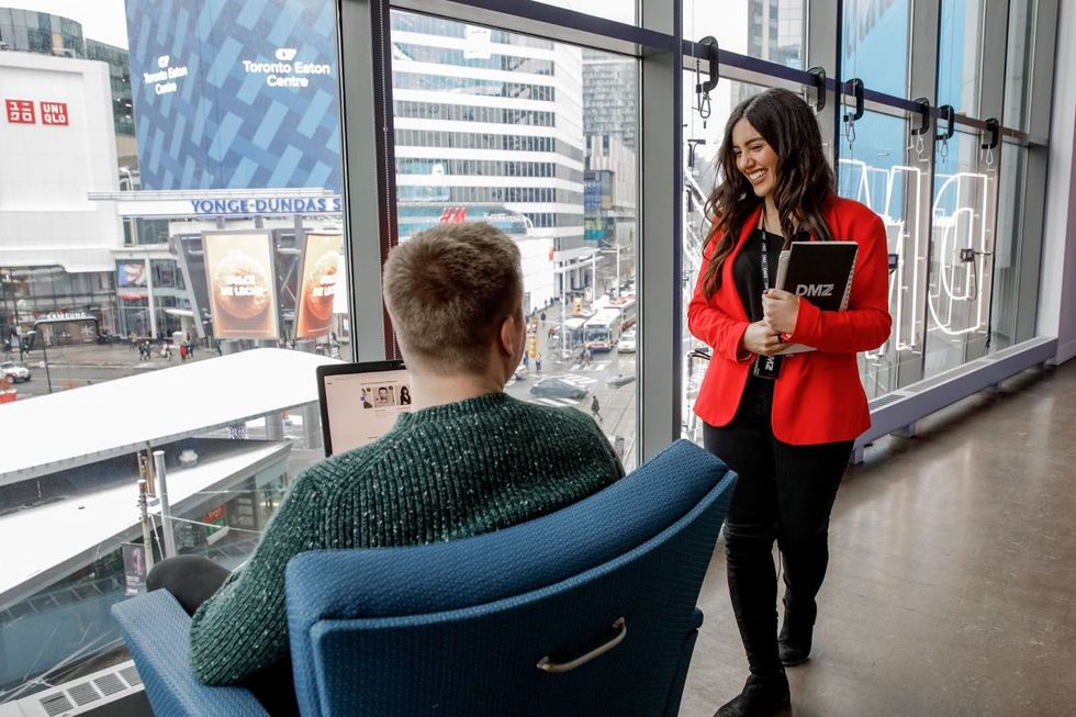 Two people talking at DMZ with views of Yonge-Dundas through the large windows.