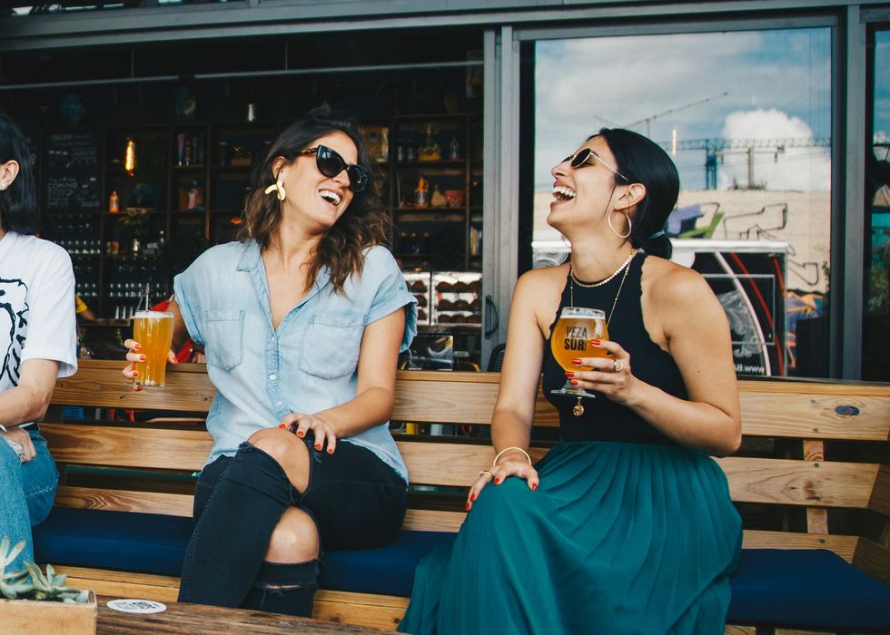 Two smiling women sit on a wooden bench, each with a drink in one hand.