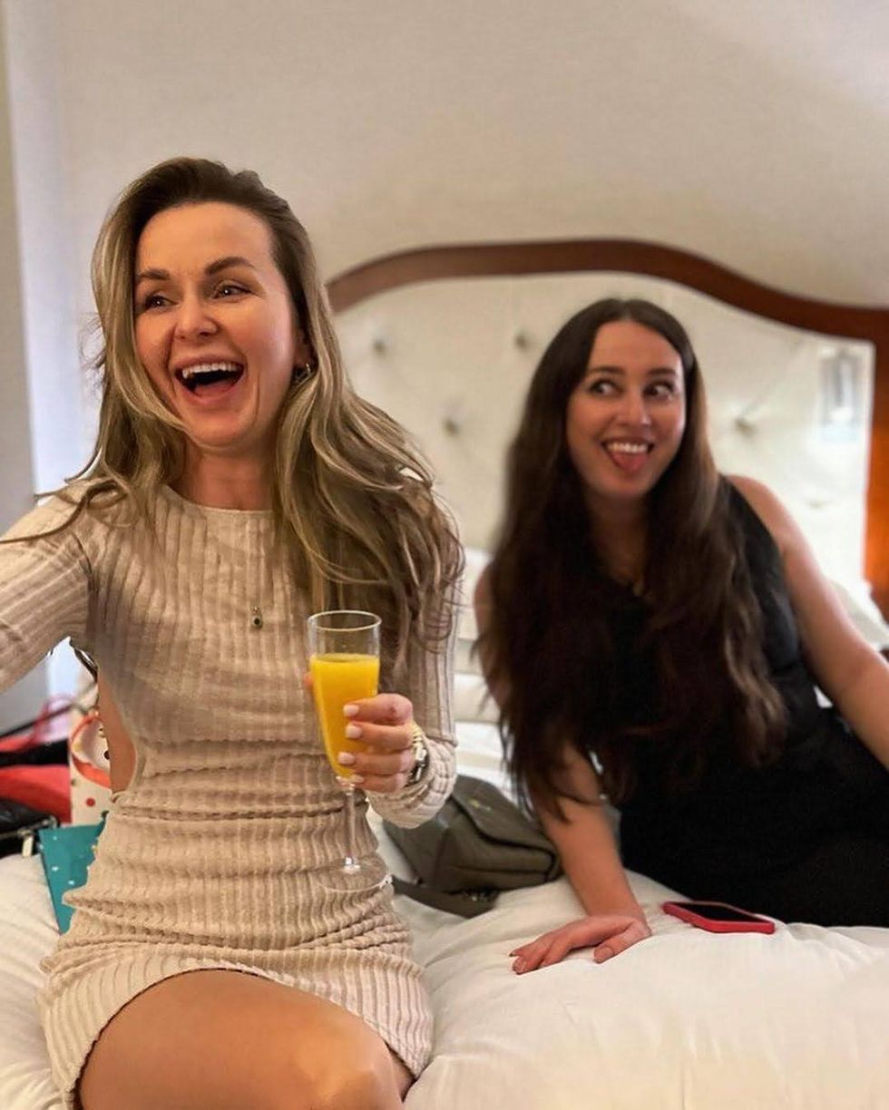 Two women laugh together while sitting on a hotel bed.