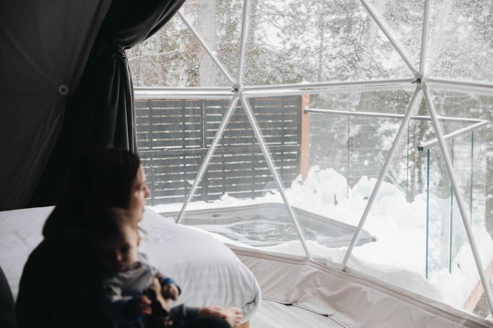 Un\u00b7e client\u00b7e regarde par la fen\u00eatre d\u2019un d\u00f4me vers un spa et la for\u00eat enneig\u00e9e.