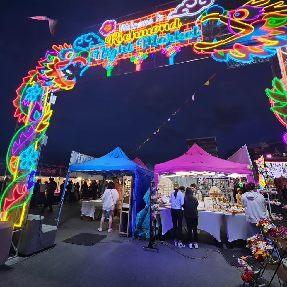 Under a neon sign reading "Richmond Night Market," customers browse market stalls selling crafts and flowers. 