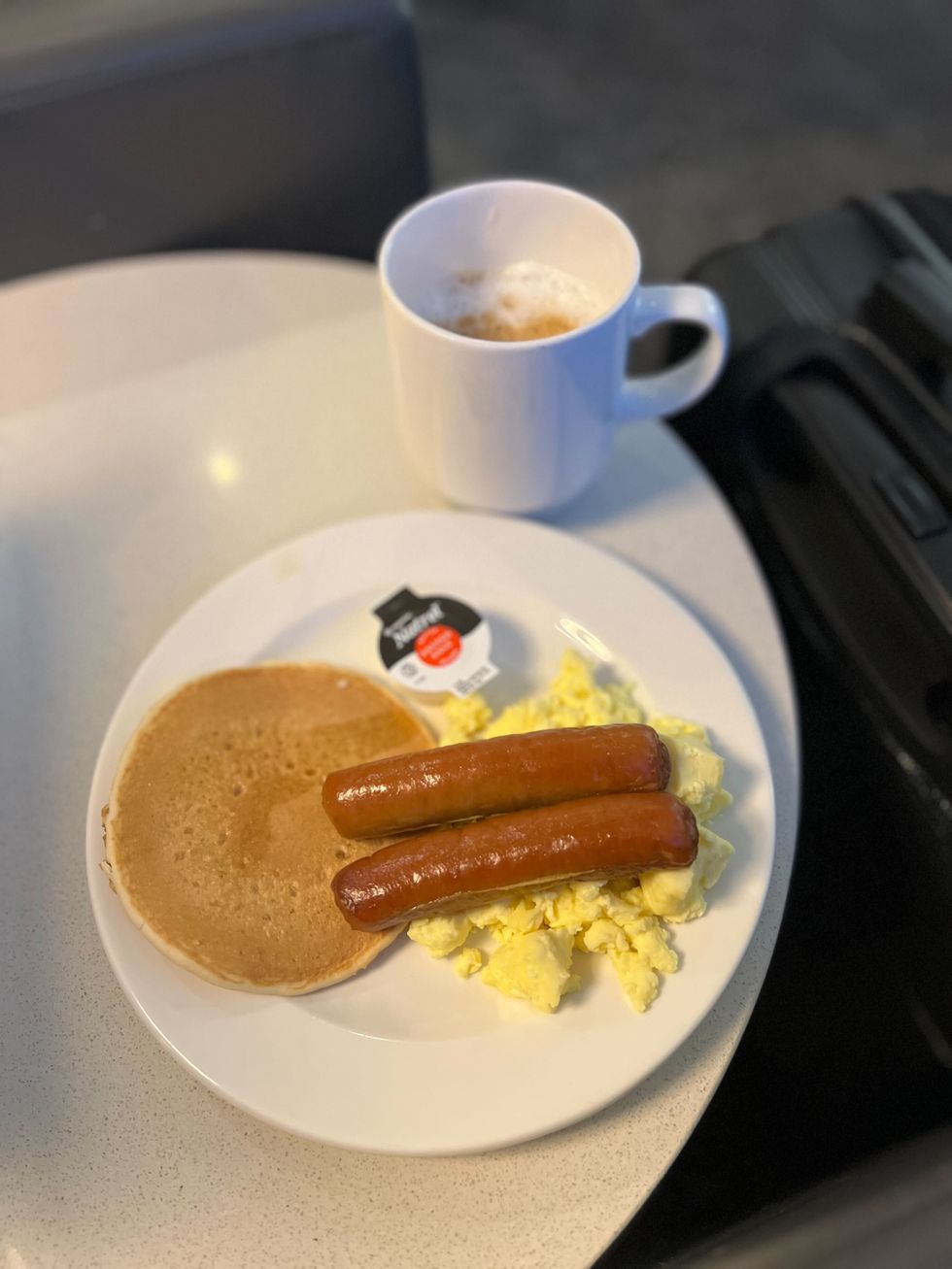 Une assiette avec des oeufs brouill\u00e9es, deux saucisses et une petite cr\u00eape \u00e0 c\u00f4t\u00e9 d'une tasse de caf\u00e9.