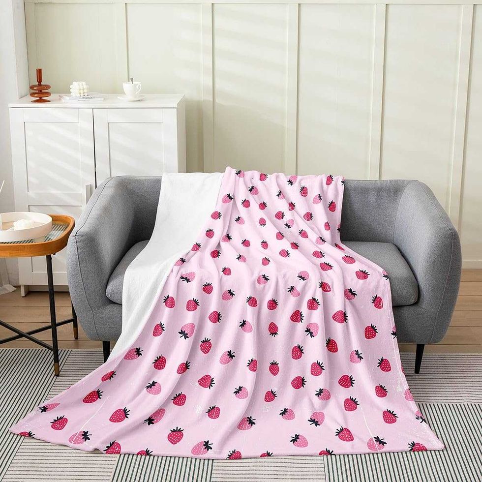 Une couverture en flanelle rose orn\u00e9e de petites fraises rouges est d\u00e9pos\u00e9e sur un canap\u00e9 gris dans un salon lumineux.