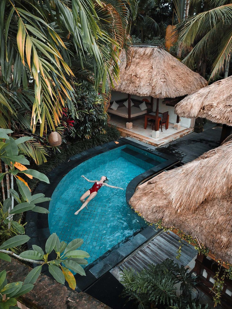 Une femme flotte sur le dos dans une piscine entoure d'arbres tropicaux.