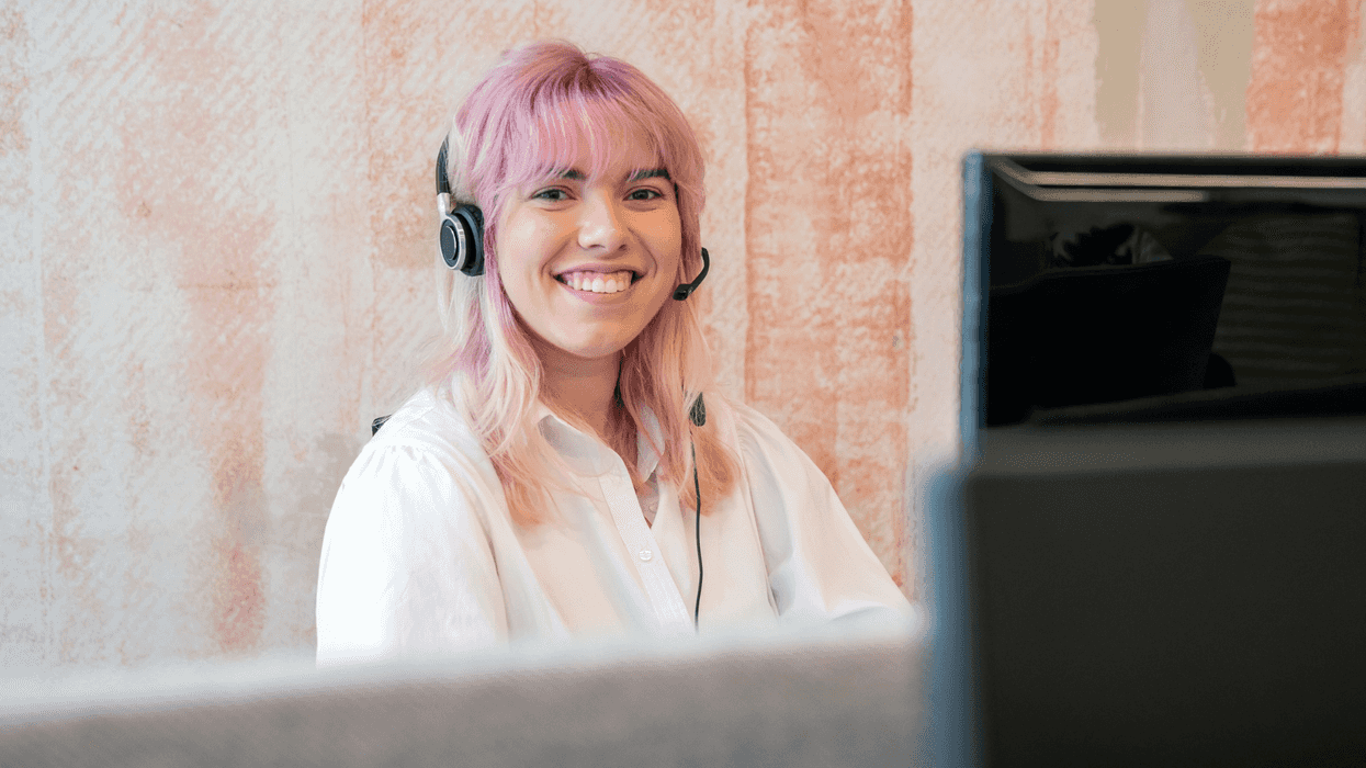 Une jeune femme aux cheveux roses portant un casque d’écoute sourit à son poste de travail.