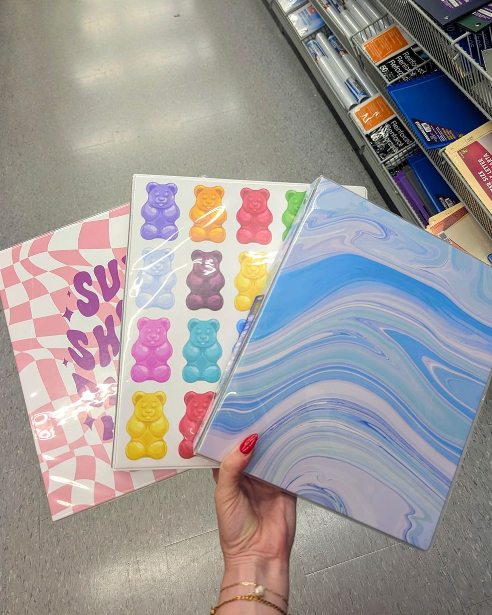 Une main qui tient 3 cartables \u00e0 motifs color\u00e9s dans la rang\u00e9e des fournitures scolaires chez Dollarama.