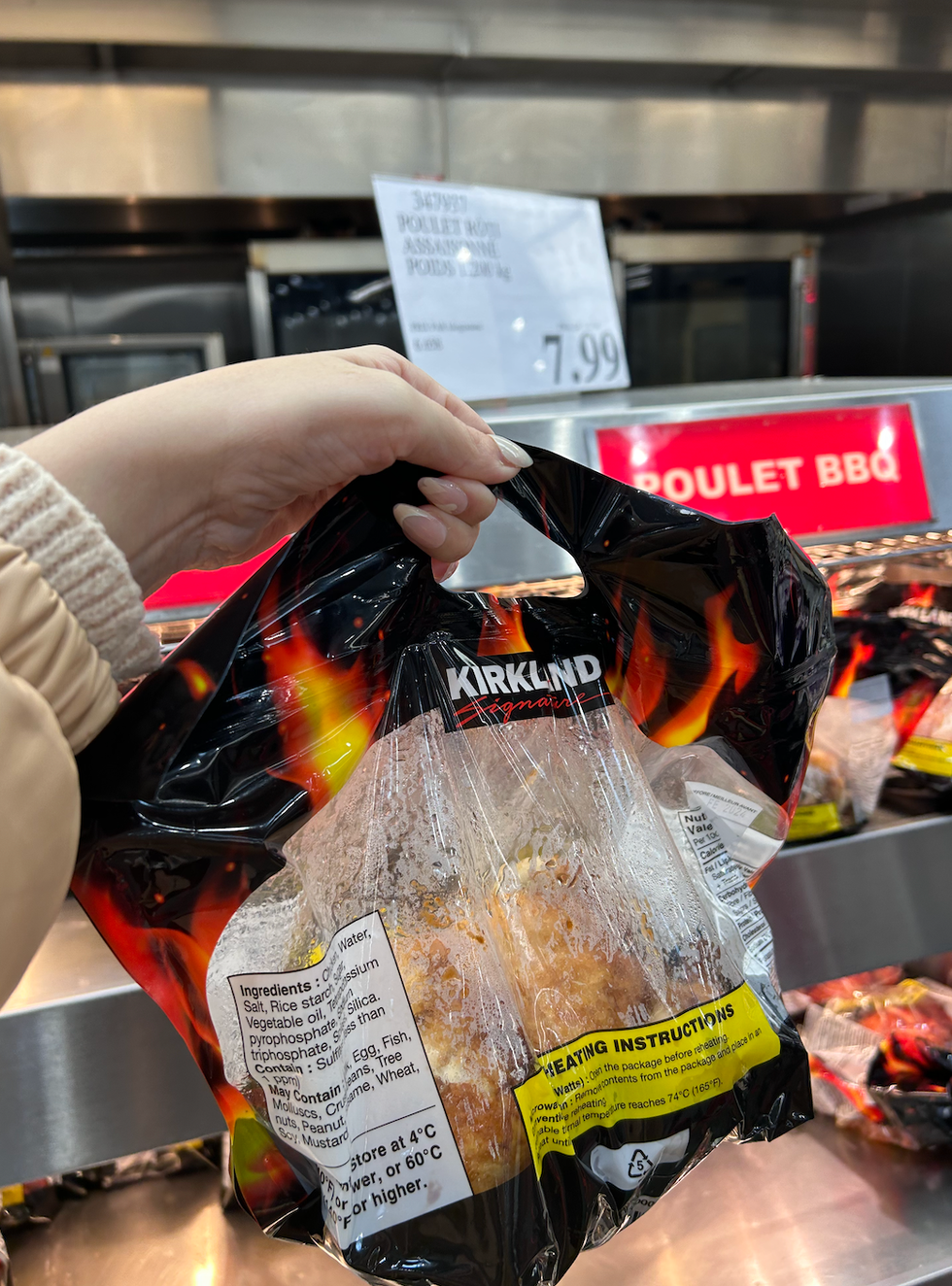 Une main tenant un sac de poulet r\u00f4ti Kirklanf Signature devant le pr\u00e9sentoir chauff\u00e9 du Costco.