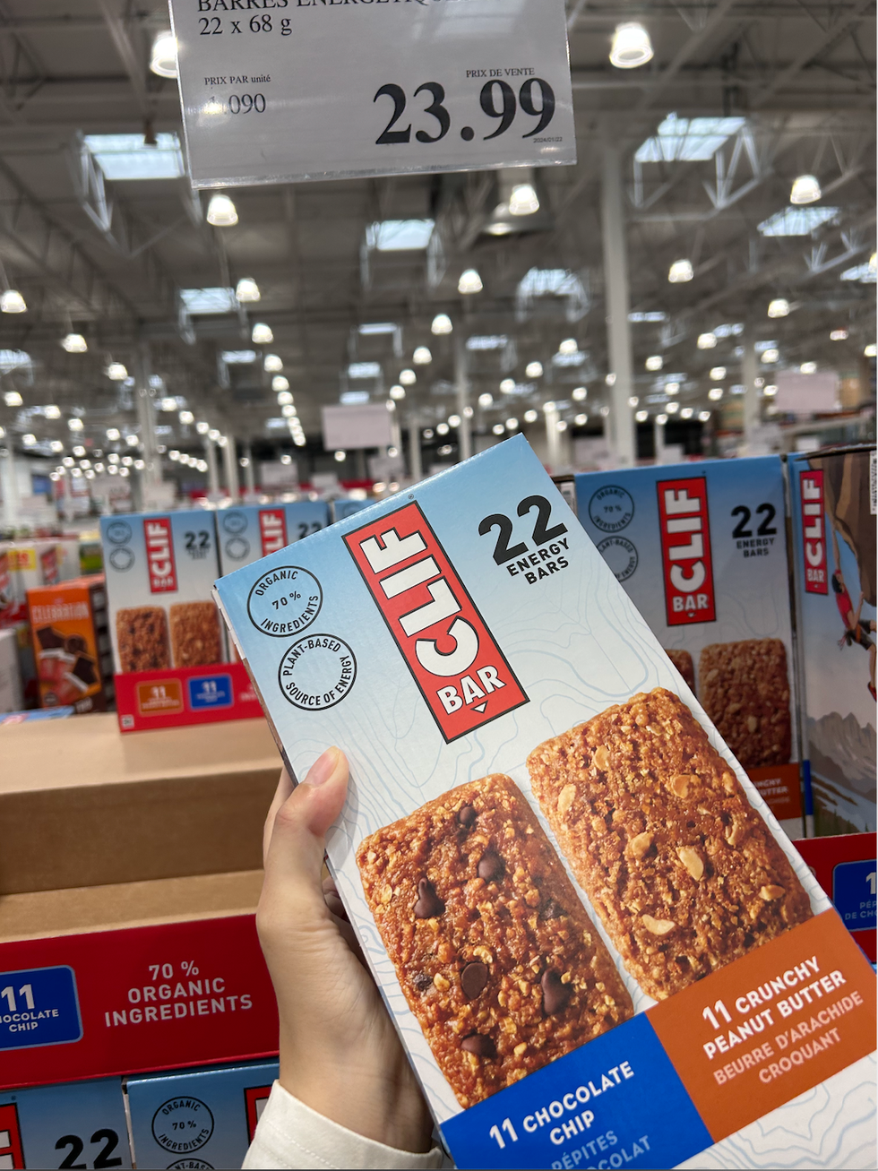 Une main tenant une boite de barres \u00e9nerg\u00e9tiques Clif Bar au p\u00e9pites de chocolat et au beurre d'arachide croquant devant un \u00e9talage du Costco.