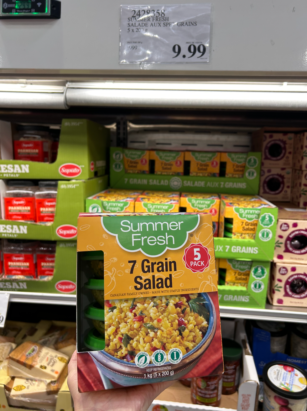 Une main tenant une boite de salades aux sept grains de Summer Fresh, devant un pr\u00e9sentoir r\u00e9frig\u00e9r\u00e9 du Costco.