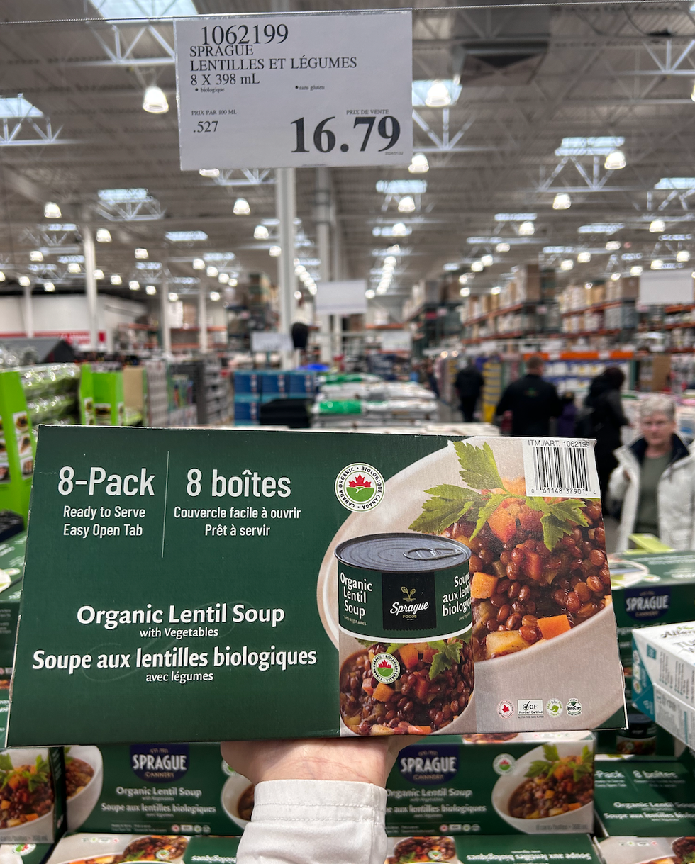 Une main tenant une boite des soupes aux lentilles et l\u00e9gumes de la marque Sprague devant un \u00e9talage du Costco.