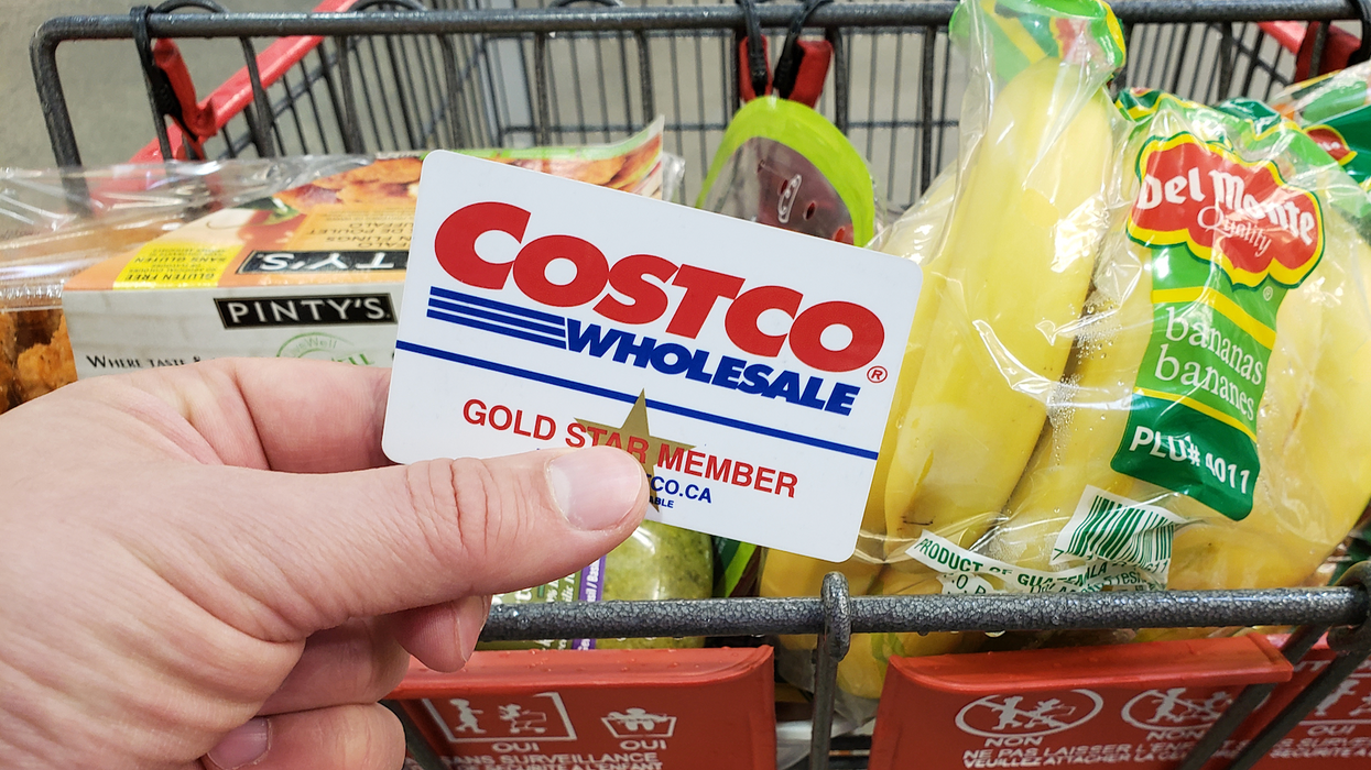Une main tenant une carte de membre du Costco devant un panier rempli d'aliments.