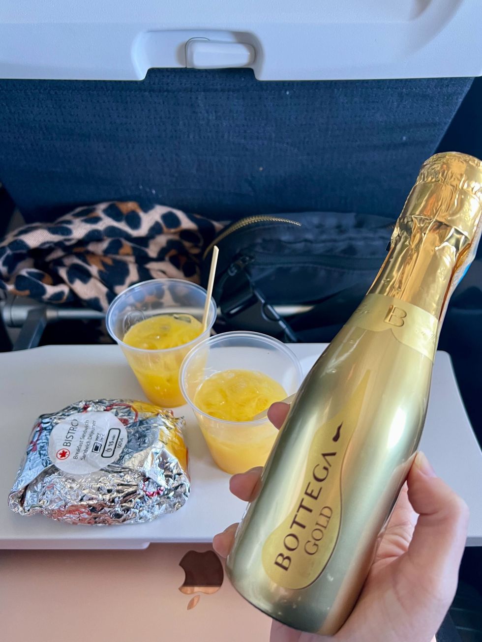 Une main tient une petite bouteille de prosecco devant deux jus d'orange et un sandwich emball\u00e9.