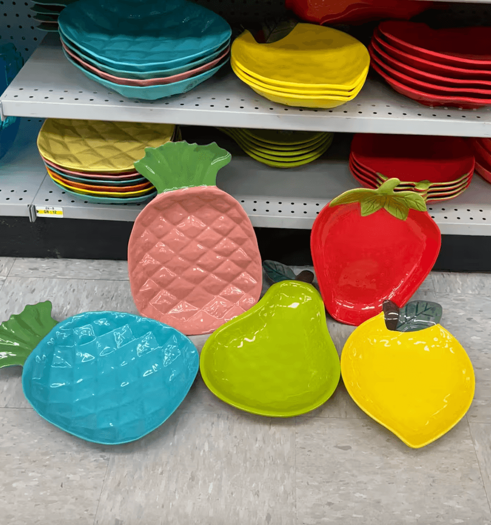 Une s\u00e9lection d'assiettes et de plateaux color\u00e9s en forme d'ananas, de citron, de fraise, de poire et de pomme, pr\u00e9sent\u00e9s sur une tablette de magasin.