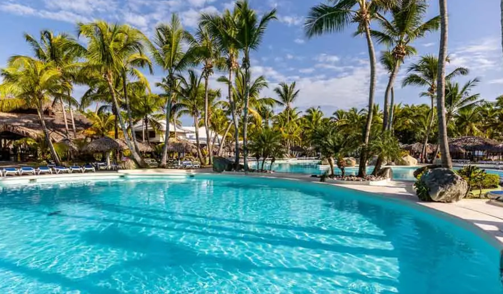 Vaste piscine turquoise entour\u00e9e de palmiers, de chaises longues et de toits de chaume.