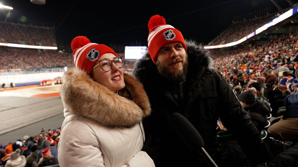 Vitoria and Mike stand in Commonwealth Stadium with the 2023 Tim Hortons NHL Heritage Classic ice behind them responding to questions from an interviewer who is not in frame. 