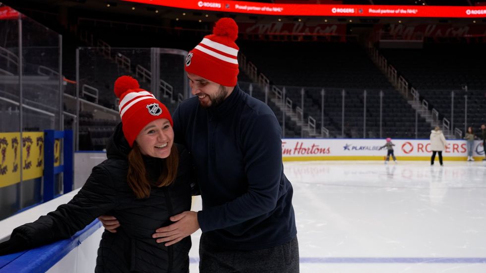 Vladimir and Zhanna wearing red NHL tuques try ice skating. Zhanna smils as she holds the edge of the rink with one hand, the other arm is around Vladimir who smiles are her while holding her up. 