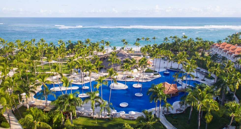 Vue a\u00e9rienne d\u2019une immense piscine bleu profond entour\u00e9e d\u2019une multitude de palmiers et de sentiers sinueux, avec la mer turquoise en arri\u00e8re-plan.