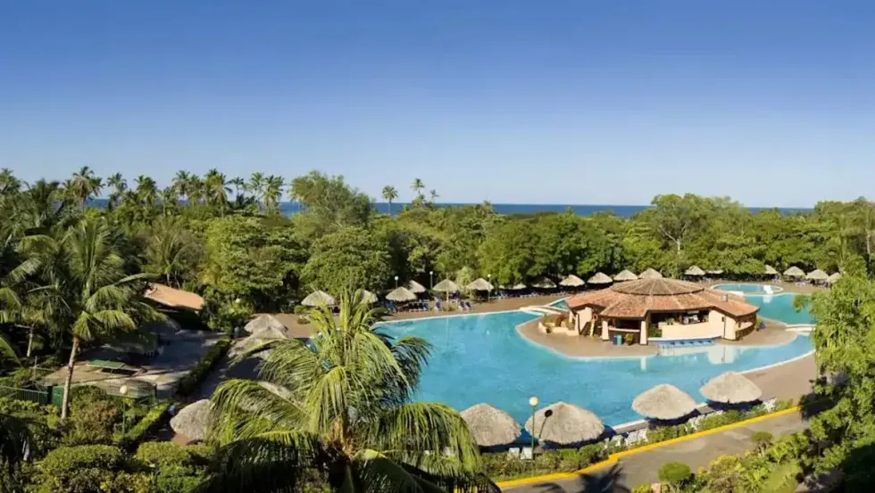 Vue a\u00e9rienne d\u2019une immense piscine entour\u00e9e de palmiers et de jardins tropicaux, avec en son centre un bar circulaire au toit de chaume.