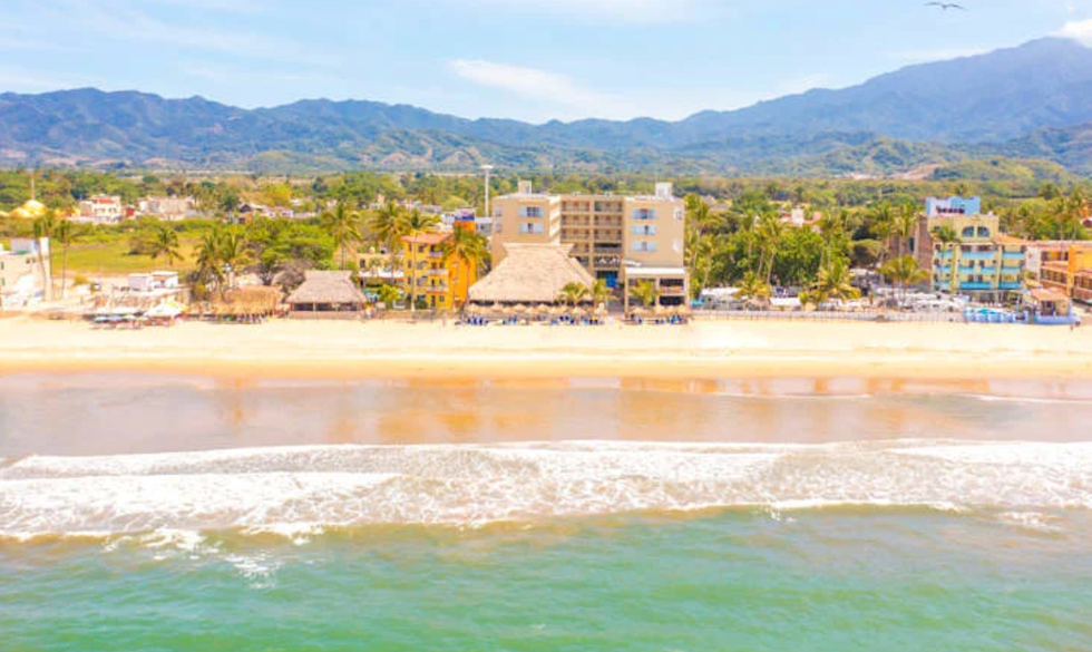 Vue de la plage devant le Decameron Isla Coral Guayabitos, bord\u00e9e par la mer et les montagnes verdoyantes au loin.
