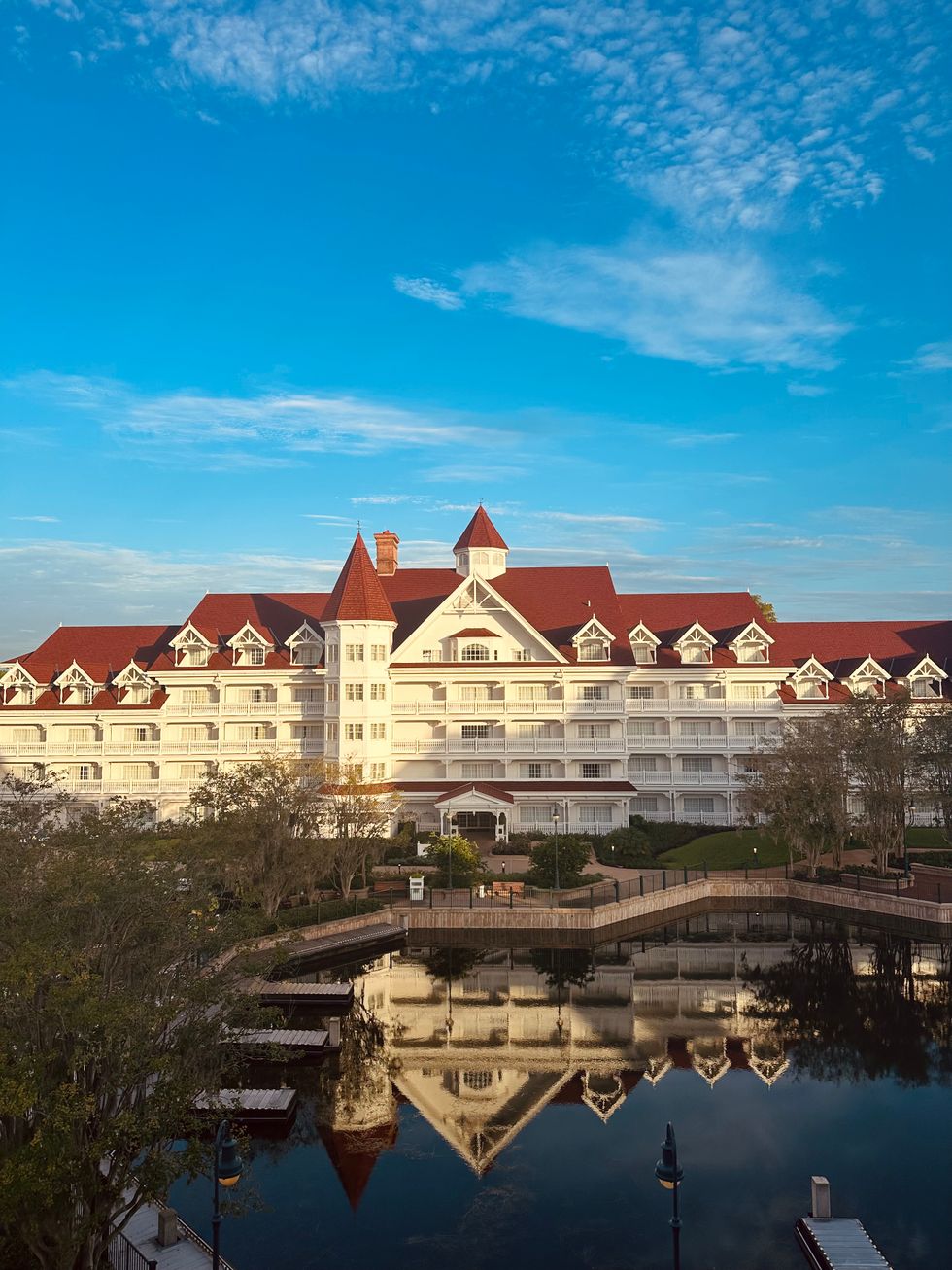 Wide shot of Disney's Grand Floridian Resort & Spa reflected in the still waters of the Seven Seas Lagoon at sunrise, with red rooftops and white Victorian architecture against a blue sky.