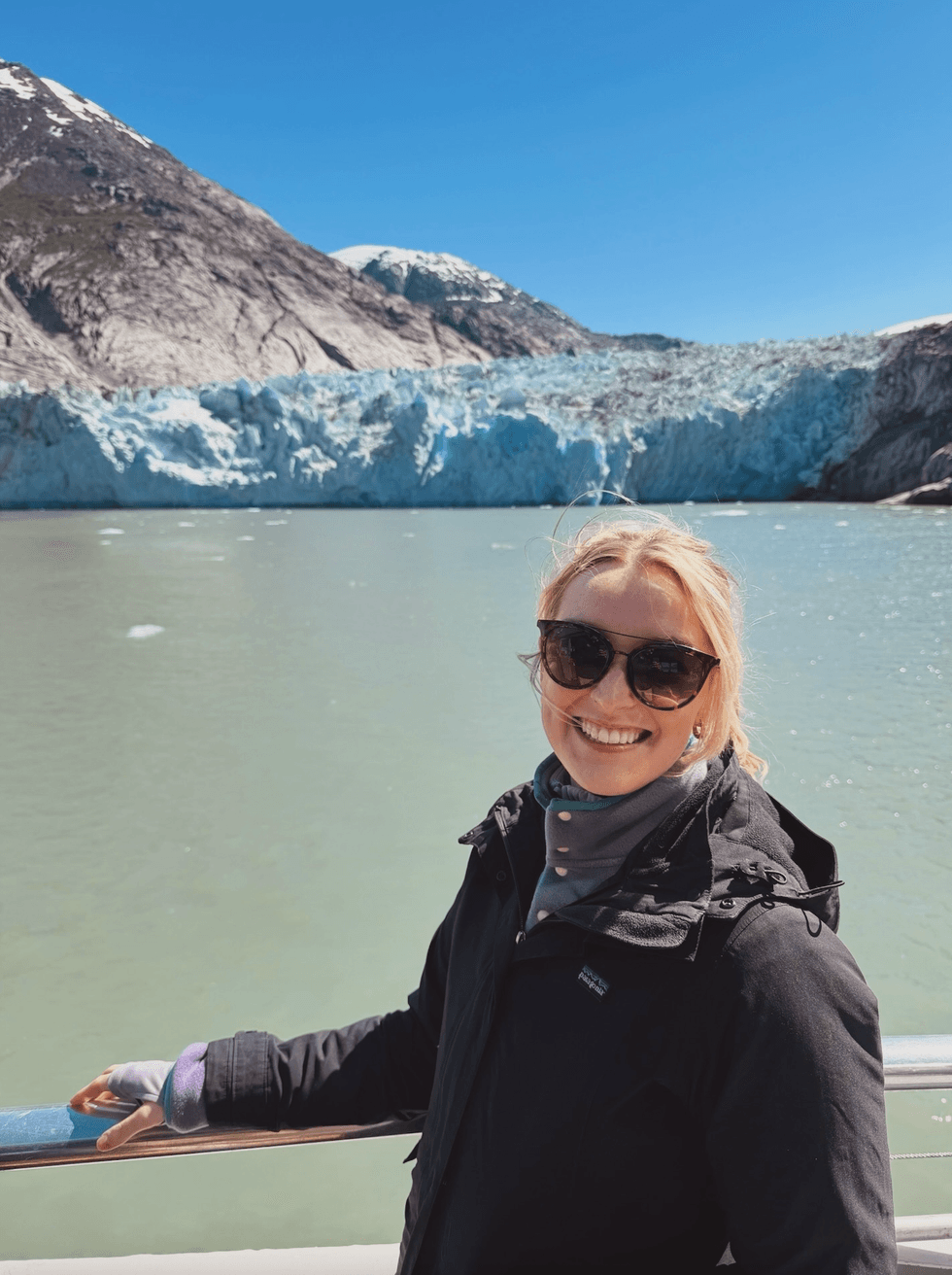 Woman in sunglasses and a black jacket smiling in front of a glacier and turquoise glacial water in Alaska.